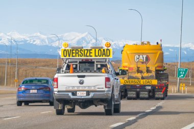 Calgary, Alberta, Kanada. 13 Nisan 2025. OVERSIZE Yük İşaretli beyaz bir pikap, yine OVERSIZE Yük damgalı büyük sarı bir CAT aracını, dağlarla çevrili bir otoyolda,.