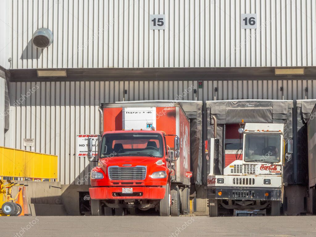 Calgary, Alberta, Canada. Apr 13, 2025. Two delivery trucks, one red and one white with the Coca-Cola logo, are parked at loading docks of a commercial building.
