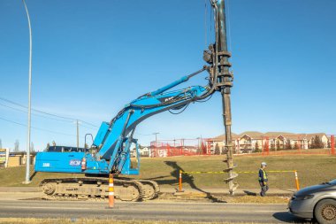 Calgary, Alberta, Kanada. 14 Nisan 2025. Mavi kazık teçhizatı bir yolun yanında zemini deliyor ve güvenlik ekipmanları takibi yapan bir işçi var. Evler arka planda açık bir gökyüzünün altında..