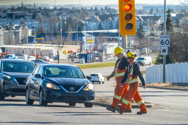 Calgary, Alberta, Kanada. 14 Nisan 2025. Turuncu takım elbiseli ve sarı şapkalı iki inşaat işçisi yerleşim alanındaki kırmızı ışıkta karşıdan karşıya geçiyor. Arabalar durdu..
