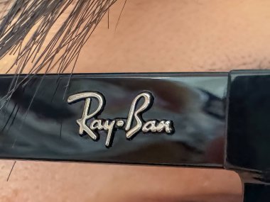 Calgary, Alberta, Kanada. 29 Nisan 2025. Işıl ışıl siyah bir çerçevede gümüş harflerle Ray-Ban logosunun yakın çekimi, muhtemelen güneş gözlüğü, bulanık bir arka plan ve en üstte görünen saçlar.