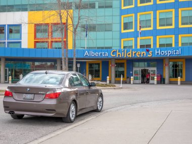 Calgary, Alberta, Kanada. 25 Nisan 2025. Kahverengi bir BMW 530i sedan Alberta Çocuk Hastanesi 'nin önüne park edilmiş, mavi, sarı ve gri aksanlı modern bir bina..