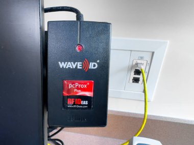 Calgary, Alberta, Kanada. 24 Nisan 2025. Bir siyah dalga ID pcProx Plus RFID okuyucusu RF IDeas tarafından bir monitöre monte edildi. Sarı bir kablo onu yakındaki bir duvar prizine Ethernet portuyla bağlıyor..