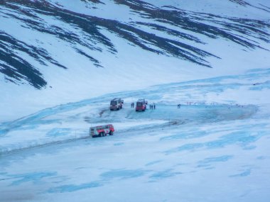 Jasper, Alberta, Kanada. 5 Mayıs 2025. Karlı, buzlu Athabasca buzulunda üç Kanadalı Icefield Ice Explorers tur otobüsü. İnsanlar otobüslerin yanında görünür ve dağlar arka planı oluşturur..