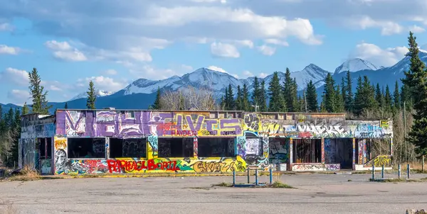 Canmore, Alberta, Kanada. 5 Mayıs 2025. Terk edilmiş, grafiti kaplı bir bina dağınık bulutlu mavi bir gökyüzünün altında duruyor. Arkaplanı karla kaplı dağlar ve yemyeşil ağaçlar oluşturuyor..