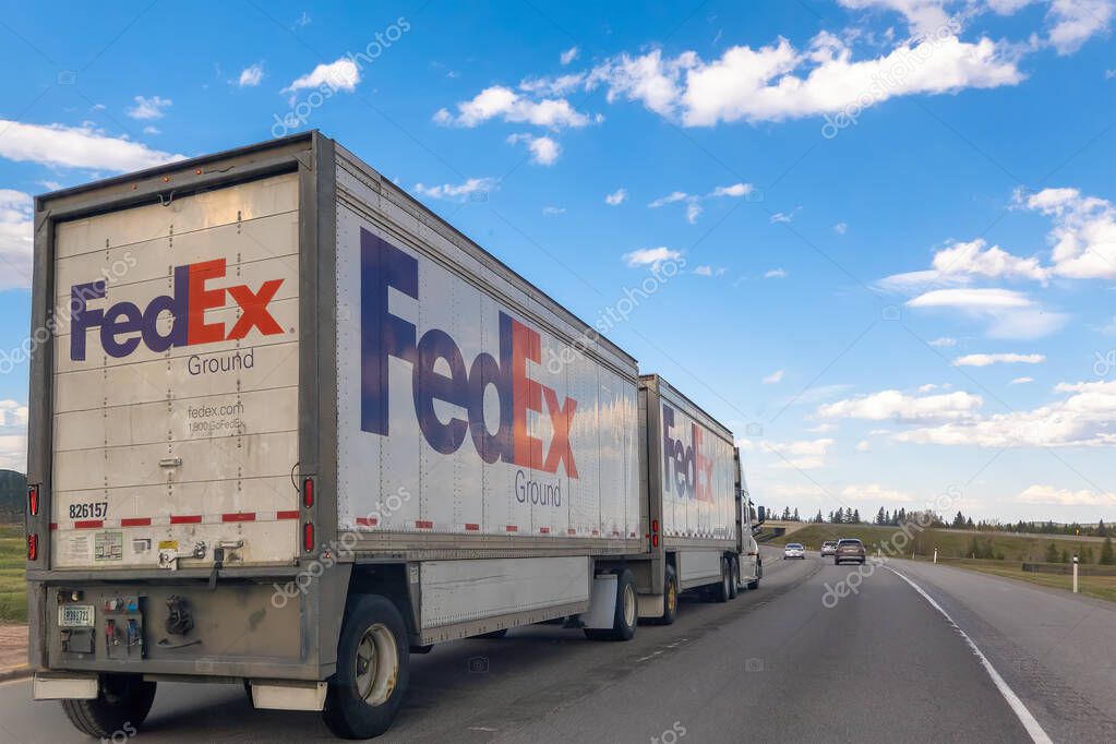 Canmore, Alberta, Canada. May 5, 2025. A white FedEx Ground double trailer truck drives on a highway under a partly cloudy sky. Other vehicles are visible in the distance.