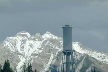 Banff, Alberta, Kanada. 9 Mayıs 2025. Bir su kulesi, bulutlu bir gökyüzünün altında karla kaplı görkemli bir zemin üzerinde duruyor. Endüstriyel yapı arasında bir zıtlık var..