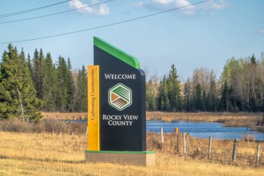 Calgary, Alberta, Kanada. 9 Mayıs 2025. Rocky View İlçesi için yol kenarında bir hoş geldin tabelası durgun bir göletin ve parçalı bulutlu bir gökyüzünün altındaki yeşil ağaçların zeminine çarpıyor..