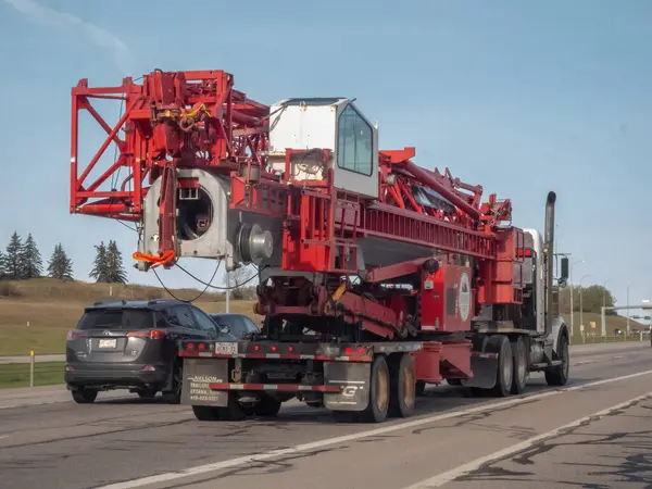 Calgary, Alberta, Kanada. 9 Mayıs 2025. Büyük bir kırmızı vinç servis platformu parçalı bulutlu bir gökyüzünün altında bir otoyol boyunca düz bir kamyonla taşınıyor..