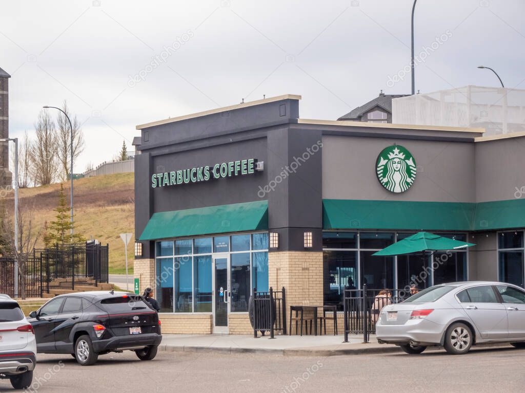 Calgary, Alberta, Canada. May 9, 2025. A Starbucks Coffee shop, presents its familiar green and white branding on a modern building under a cloudy sky, with parked cars nearby.