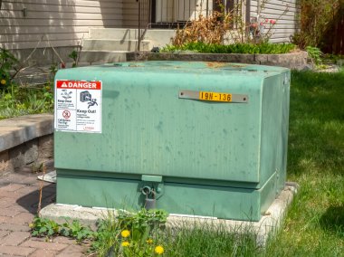 Calgary, Alberta, Kanada. 16 Mayıs 2025. Bir banliyö evinin ve yeşil çimlerin arka planına uyarı etiketleriyle süslenmiş, beton bir zeminde elektrikli yeşil bir hizmet kutusu bulunuyor..