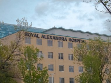 Edmonton, Alberta, Kanada. 24 Mayıs 2025. Kraliyet Alexandra Hastanesi, yumuşak, bulutlu bir gökyüzünün altında yemyeşil ağaçlar tarafından çerçevelenmiş ılık yüzüyle sağlık feneri olarak duruyor..