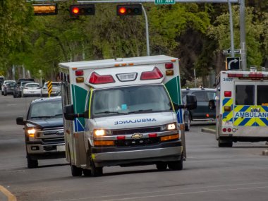 Edmonton, Alberta, Kanada. 24 Mayıs 2025. Bir Alberta Sağlık Servisi ambulansı yeşil ağaçlar ve şehir trafiği arasında şehir caddesinde hızla ilerliyor..