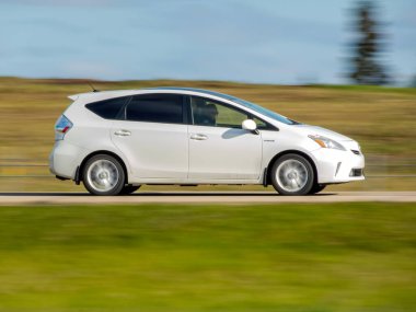 Edmonton, Alberta, Kanada. 24 Mayıs 2025. Beyaz bir Toyota Prius V, 2013 model, hibrit tasarımını gösteren bulanık bir arka planı olan bir yol boyunca sürüyor..