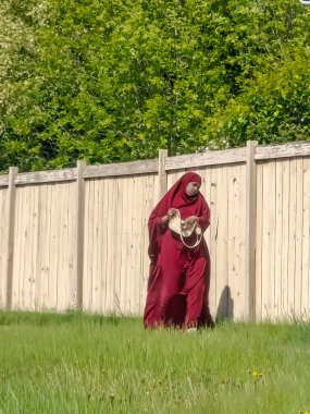 Calgary, Alberta, Kanada. 27 Mayıs 2025. Uzun kırmızı elbiseli ve başı örtülü bir kadın çimenli bir alanda yürüyor, kültürel ve dini çeşitliliği ve bireysel ifadeyi somutlaştırıyor..
