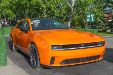 Calgary, Alberta, Kanada. 28 Haziran 2025. Modern bir kaslı araba olan parlak turuncu Dodge Charger Daytona EV, verimli yeşilliklerle çevrili, güneşin altında sergilenmektedir..