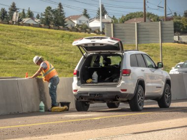 Calgary, Alberta, Kanada. 27 Haziran 2025. Güvenlik donanımlı bir inşaat işçisi, yol kenarı bakım ve altyapı bakımını yansıtan bir sprey şişeyle beton bariyeri temizliyor..
