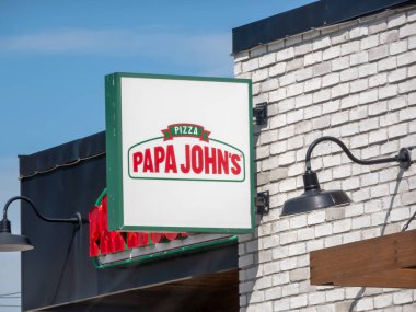 Calgary, Alberta, Canada. Aug 22, 2025. A close-up of a Papa John's Pizza sign on a white brick building, and a blue sky in the background.