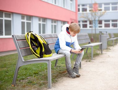 Okul çocuğu Sırt çantasıyla Bench 'te oturuyor ve telefona bakıyor. Yüksek kalite fotoğraf