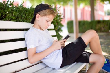 Güneşli bir günde ahşap bir bankta oturan genç, akıllı telefonunu kullanırken odaklanmış görünüyor. Yüksek kaliteli fotoğraf..