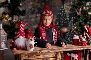 Şirin çocuk, sarışın çocuk, tahta bir standın üzerinde duruyor, Noel için süslenmiş, sıcak çikolata içiyor ve kurabiye yiyor.