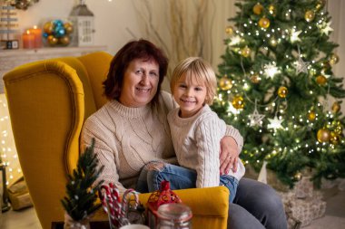 Sevimli çocuk, oğlum, Noel 'de anne ve büyükanneyle süslü bir odada sarı koltukta oturuyor.