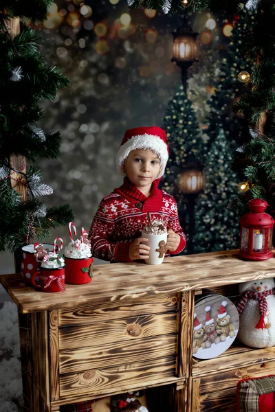 かわいい幼児の子供 ブロンドの男の子 木製のスタンドの雪の中に立って クリスマスのために飾られ 熱いチョコレートを飲んで クッキーを食べる — ストック写真