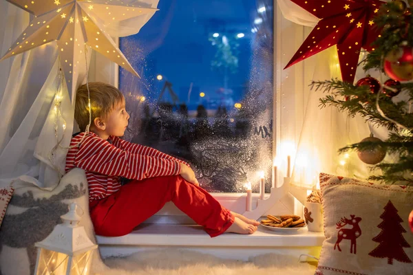 幼児の子供 かわいいブロンドの男の子 パジャマの窓の上に座って サトナ クラウスを探して クッキーを食べて 彼の周りのクリスマスライト — ストック写真