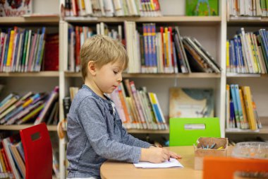 Adorable little child, boy, sitting in library, reading book and choosing what to lend, kid in book store