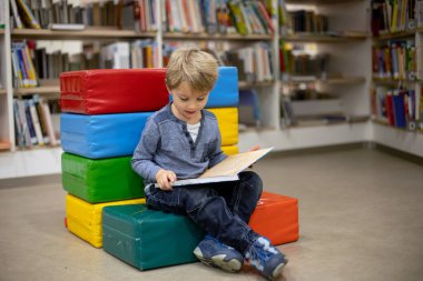 Adorable little child, boy, sitting in library, reading book and choosing what to lend, kid in book store