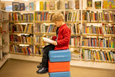 Adorable little child, boy, sitting in library, reading book and choosing what to lend, kid in book store