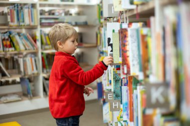 Adorable little child, boy, sitting in library, reading book and choosing what to lend, kid in book store