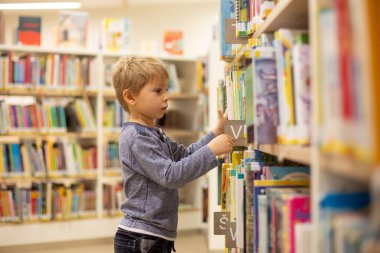 Adorable little child, boy, sitting in library, reading book and choosing what to lend, kid in book store