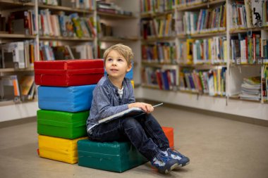 Adorable little child, boy, sitting in library, reading book and choosing what to lend, kid in book store
