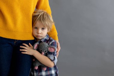 Üzgün küçük çocuk, sarışın çocuk, evde annesine sarılıyor, oyuncak ayıyı tutuyor.