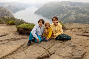 Family, enjoying the hike to Preikestolen, the Pulpit Rock in Lysebotn, Norway on a rainy day, toddler climbing with his pet dog the one of the most scenic fjords in Norway