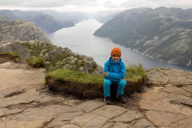 Family, enjoying the hike to Preikestolen, the Pulpit Rock in Lysebotn, Norway on a rainy day, toddler climbing with his pet dog the one of the most scenic fjords in Norway