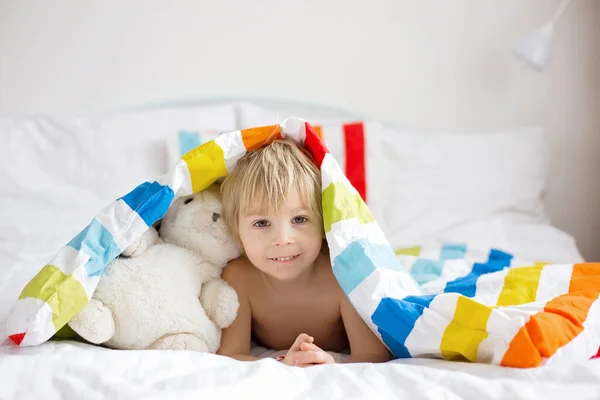 幸せな幼児の子供 カラフルなバスローブを着たブロンドの男の子 バスの後にぬいぐるみでベッドに座って カラフルな寝具 — ストック写真