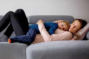 Anne hasta bir çocuğu kucağında tutuyor, ateşli bir şekilde kanepede uzanıyor, burnu ve ateşi akıyor, dinleniyor.