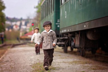 İki çocuk, eski moda kıyafetler ve şapka giymiş, bavullu, tren istasyonunda.