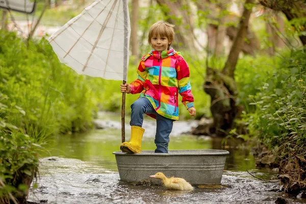 Cute child, boy in colorful jacket, playing with boat and ducks on a little river, sailing and boating. Kid having fun, childhood happiness concept