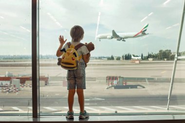 Çocuk, havaalanının penceresinden uçaklar havalanıyor ve iniyor. Tatil için uçağa binmeyi bekliyor.