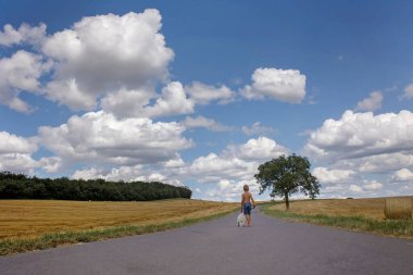 Güzel sarışın çocuk, evlat, kırsal yolda tatlı küçük köpeğiyle yürüyor. İnanılmaz manzara, bulutlu kırsal manzara, ağaç ve boş yol yaz mevsimi, yol kenarındaki saman yığını, yaz mevsimi.