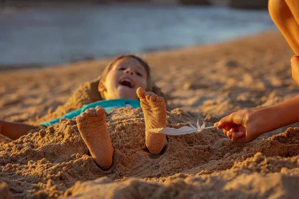Child, tickling sibling on the beach on the feet with feather, kid cover in sand, smiling, laughing, enjoying some fun