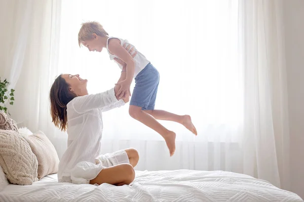 Mother, playing with her preschool child at home in bed, having fun jumping and flying