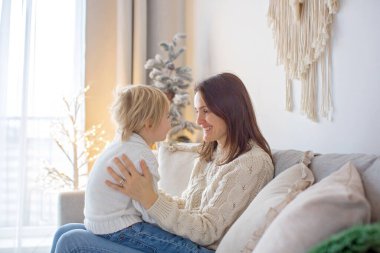 Güzel sarışın çocuk, oğlum, anneye kanepede sarılıp anneye gülümsüyor.