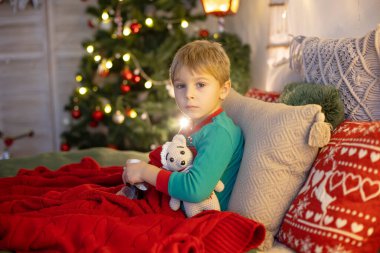 Küçük sarışın çocuk, yatağında hasta yatıyor, şurup içiyor, üzülüyor.