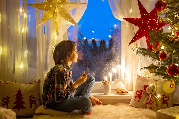 蹒跚学步的孩子 可爱的金发男孩 穿着睡衣坐在窗上 望着萨特娜 克劳斯 吃着饼干 身边点着圣诞灯 — 图库照片