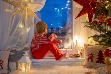 Küçük bir çocuk, tatlı sarışın çocuk, pijamayla pencerede oturuyor, Satna Claus 'a bakıyor, kurabiye yiyor, Noel ışıklarını yakıyor.