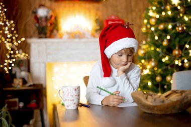 Güzel sarışın çocuk, genç okul çocuğu süslü bir evde Noel Baba 'ya mektup yazıyor süt içiyor ve kurabiye yiyor. Işıklar ve örgü oyuncaklar